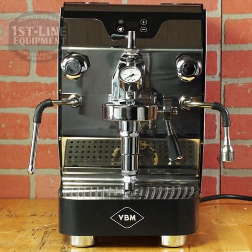 A *SOLD* SIDEWALK SALE VBM Domobar Junior Digital HX Espresso Machine w/ Flow Control (SAMPLE) (E014) rests on a wooden counter before a red brick wall, featuring a pressure gauge, two steam wands, and a chrome group head. © 2025 1st-line.com