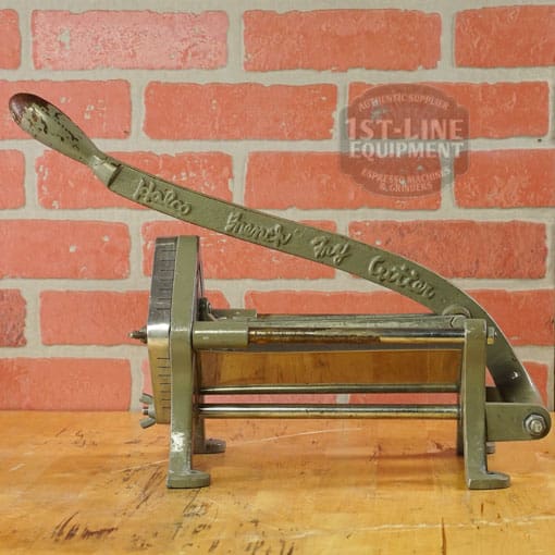 A vintage manual coffee grinder with a long handle and metal frame sits on a wooden surface in front of a red brick wall—ideal for your next SIDEWALK SALE or to pair with the Halco French Fry Cutter Maker (E110). © 2025 1st-line.com