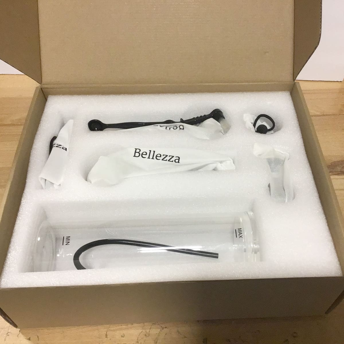 An open cardboard packing box with foam padding contains Bellezza glass and metal parts, including a clear glass tube, black metal wand, and small accessories, all neatly arranged and partially wrapped in paper. © 2025 1st-line.com