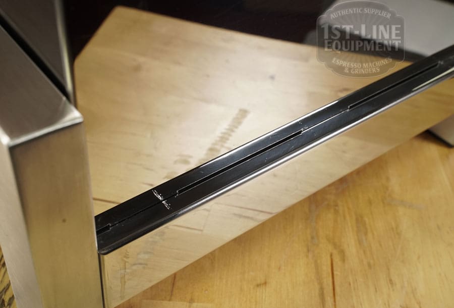 Close-up of a LIKE NEW - Elektra Verve Dual Boiler Espresso Machine (C104) featuring a black edge strip with visible metal scratches; a wooden table and partial logo are reflected in the background. © 2025 1st-line.com