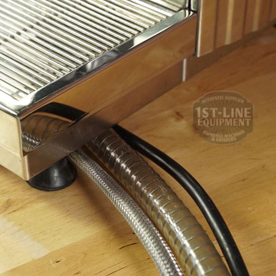 Close-up of the bottom corner of a SOLD SIDEWALK SALE - Fiorenzato Bricoletta Electronic Direct Connect HX Espresso Machine (E011) on wood, showing two hoses—one braided metal, one black plastic—emerging from underneath. © 2025 1st-line.com