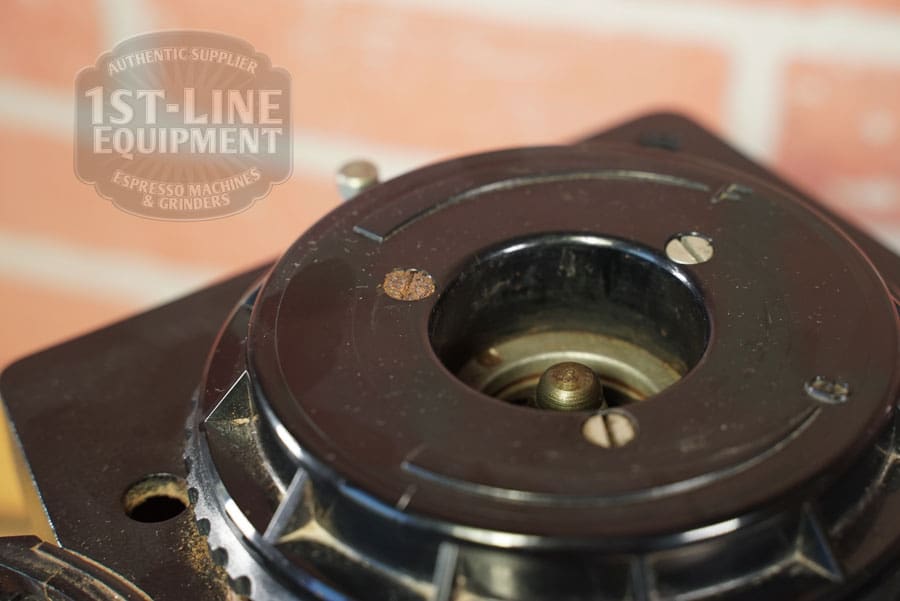 Close-up of the SIDEWALK SALE - Cunill Doser Grinder (E012) burr holder housing with rust spots, set against a blurred brick wall. 1st-Line Equipment logo is watermarked in the top left corner—great for the upcoming sale!. © 2025 1st-line.com