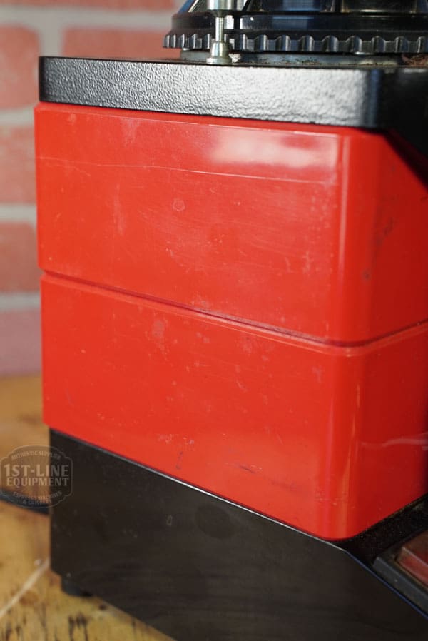 Close-up of the SIDEWALK SALE - Cunill Doser Grinder (E012) in red and black, featuring a boxy design, smooth surfaces, and a top dial, set against a brick wall background. © 2025 1st-line.com