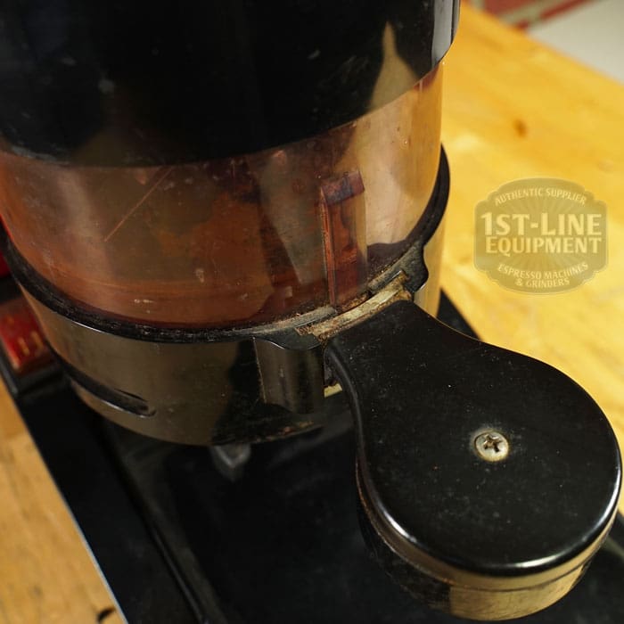 Close-up of the SIDEWALK SALE - Cunill Doser Grinder (E012) with coffee grounds inside, set on a wooden surface. The transparent hopper displays the 1st-Line Equipment logo—your next great Sidewalk Sale discovery!. © 2025 1st-line.com