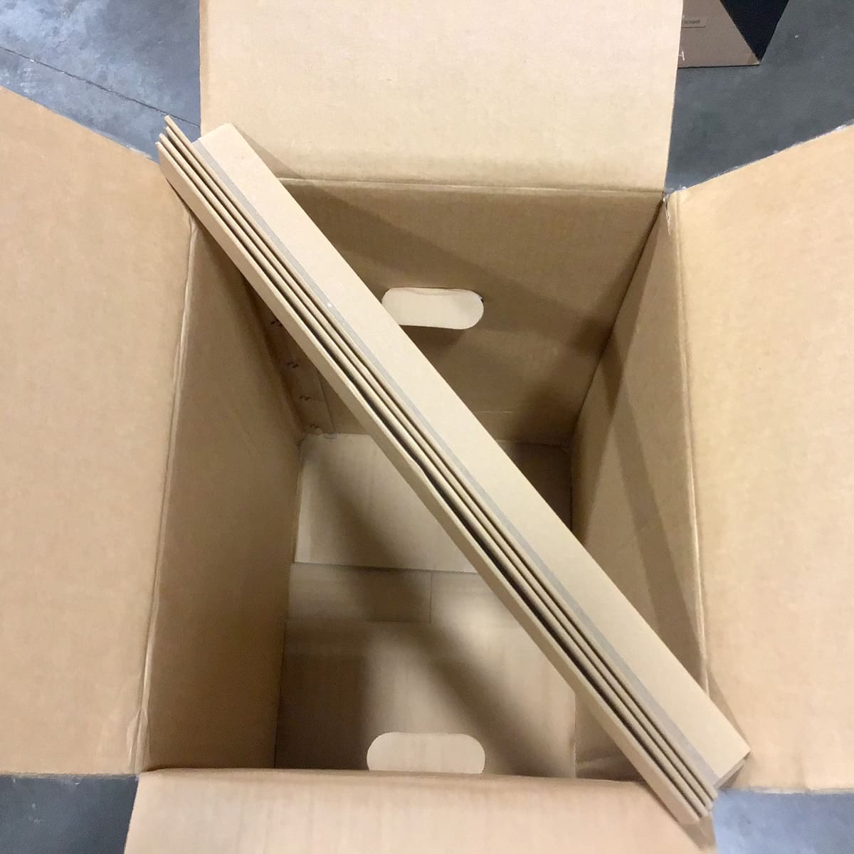 Open cardboard box with several flat, unfolded packing dividers resting diagonally inside on top of the box’s bottom flaps, placed on a concrete floor—ideal for organized packing with Bellezza simplicity. © 2025 1st-line.com