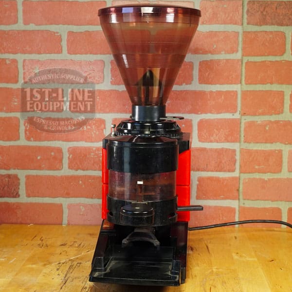 The SIDEWALK SALE - Cunill Doser Grinder (E012) features a large brown hopper and black base, set on a wooden countertop against a red brick wall, plugged in with fresh coffee grounds ready in the container. © 2025 1st-line.com