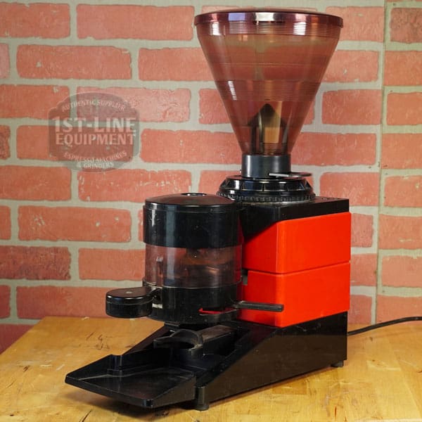 The SIDEWALK SALE - Cunill Doser Grinder (E012) in red and black sits on a wooden surface before a brick wall, featuring a large transparent hopper and front dispensing area—ideal for your next sidewalk sale. © 2025 1st-line.com