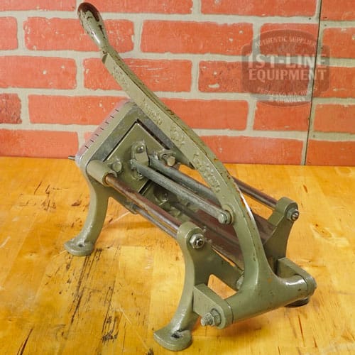 The SIDEWALK SALE - Halco French Fry Cutter Maker (E110) features a lever handle and is displayed on a wooden surface against a red brick wall—an ideal find for your next sidewalk sale. © 2025 1st-line.com