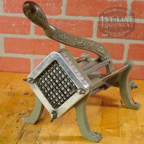 The SIDEWALK SALE - Halco French Fry Cutter Maker (E110) with lever handle rests on a wooden surface by a red brick wall, perfect for slicing potatoes into fries—an excellent choice for Sidewalk Sale shoppers. © 2025 1st-line.com