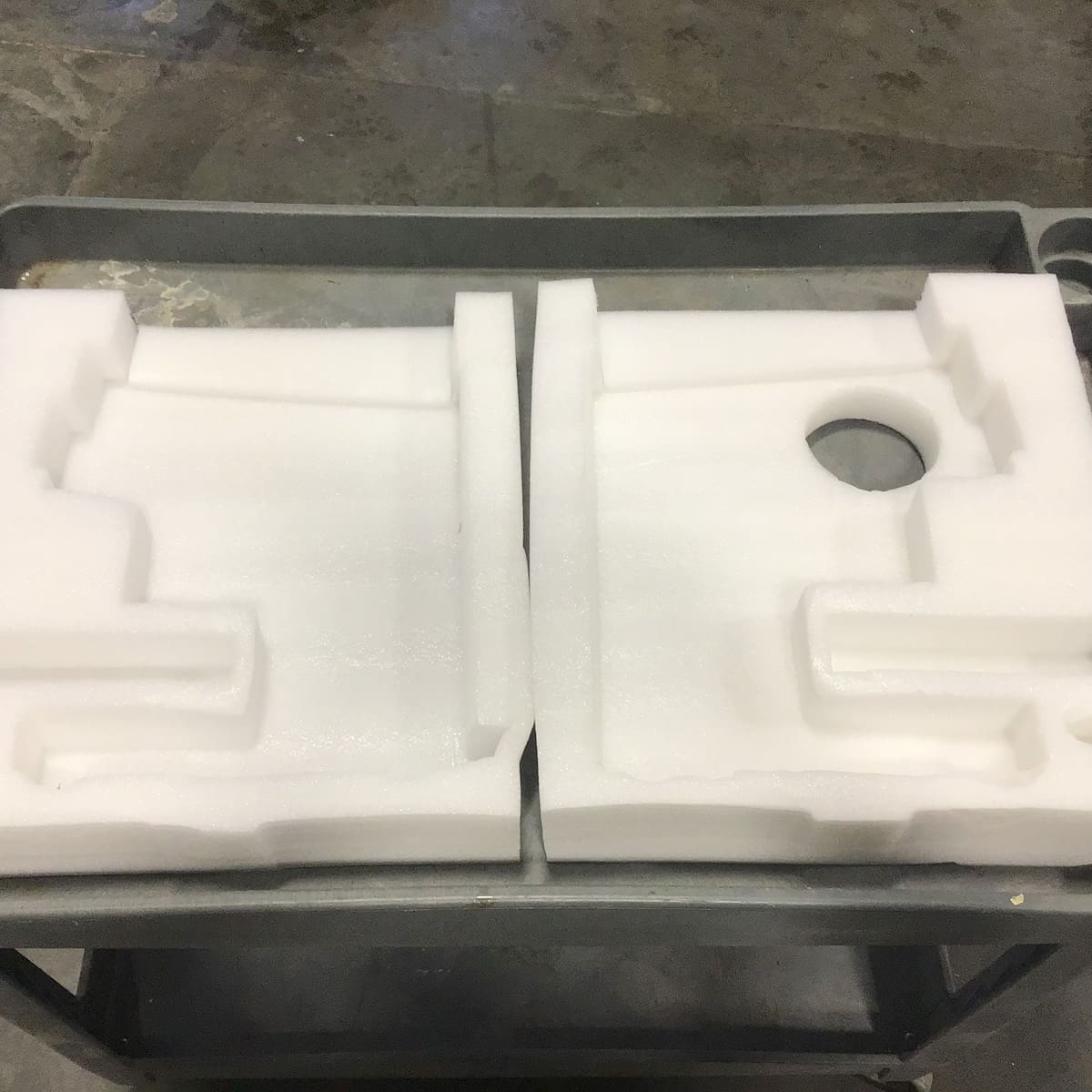 Two white foam packing inserts with custom cutouts and a circular hole in one piece are placed side by side on a gray metal cart, showcasing Bellezza-quality packing against a concrete floor background. © 2025 1st-line.com