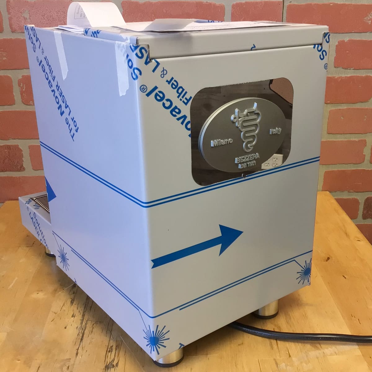 A Bezzera Unica Espresso Machine (Returned), featuring blue protective wrap and an engraved logo, is placed on a wooden table in front of a red brick wall, showing markings that indicate it is a returned item. © 2025 1st-line.com