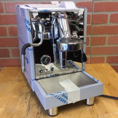 A Bezzera Unica Espresso Machine (Returned) sits on a wooden table before a red brick wall, featuring a pressure gauge, steam wand, and protective tape that indicate it may be a recently returned unit. © 2025 1st-line.com