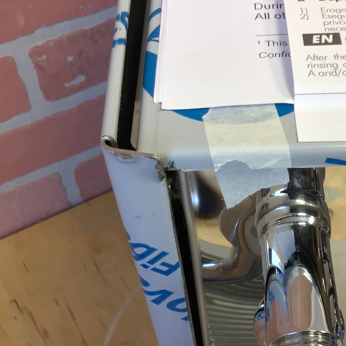 A close-up of a Bezzera Unica Espresso Machine (Returned), partially unwrapped with protective blue and white film, sitting on a wooden surface near a pink brick wall and white papers taped above. © 2025 1st-line.com