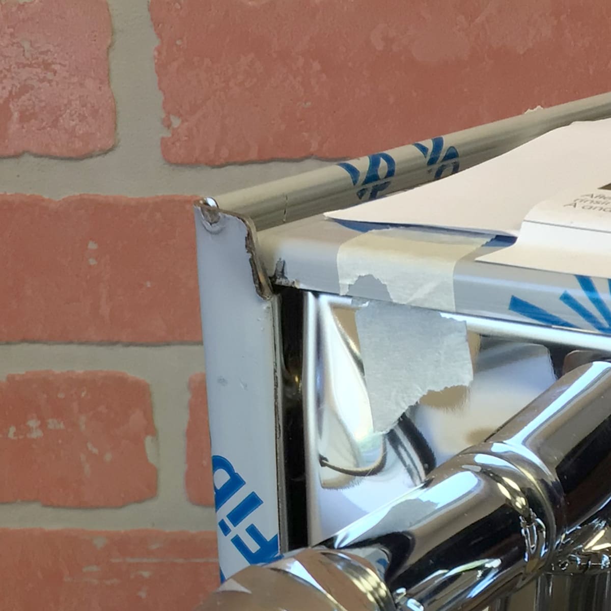 A close-up of the Bezzera Unica Espresso Machine (Returned) highlights its metal fixture with protective film peeling off and tape at the corner, set against a brick wall with light-colored mortar in the background. © 2025 1st-line.com