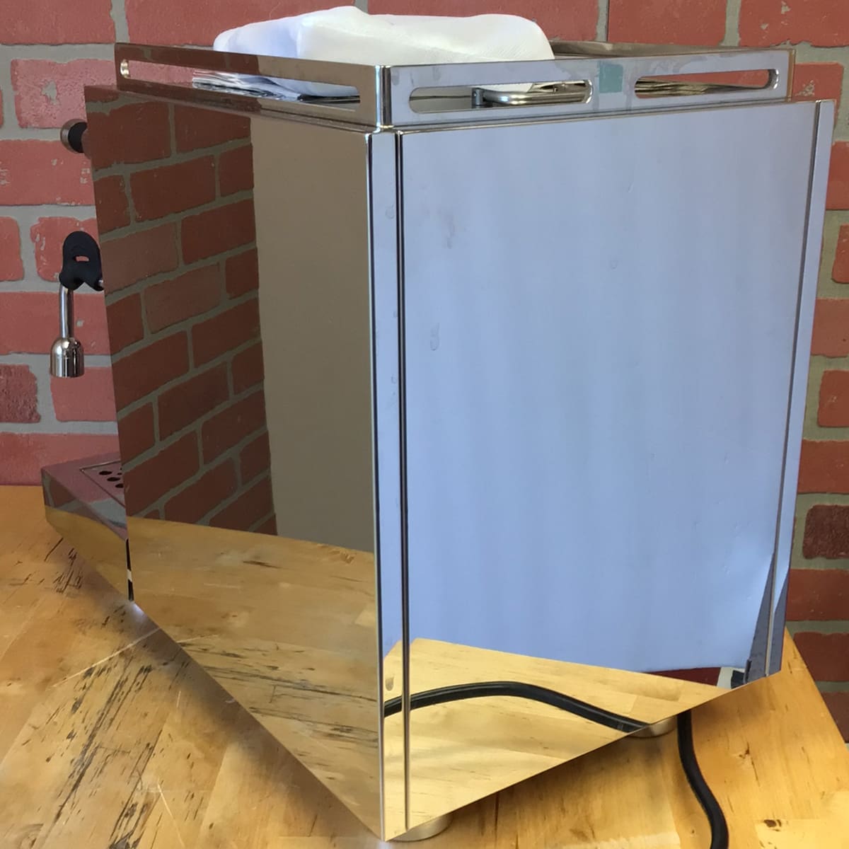 A SIDEWALK SALE - Bellezza Inizio R Leva HX Espresso Machine (USED)(E314) with a reflective side panel sits on a wooden table before a red brick wall, its power cord extending from the back. © 2025 1st-line.com