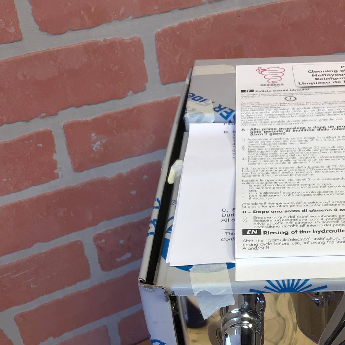 A stack of Italian and English papers rests atop a sealed box labeled Bezzera Unica Espresso Machine (Returned), placed on a chrome stand beside a red and white brick-patterned wall. © 2025 1st-line.com