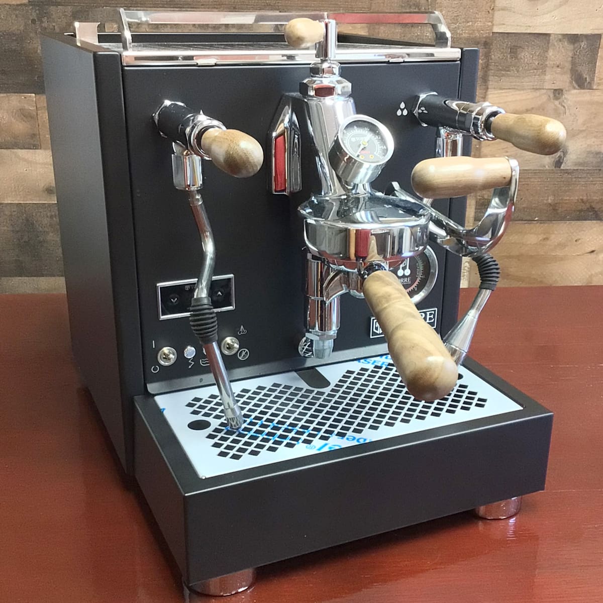 A Torre Mariuccia Dual Boiler Espresso Machine w/ Flow Control (demo) (E313) in chrome and black with wooden handles sits on a wooden surface, featuring flow control gauges, steam wands, and a drip tray against a rustic wood wall. © 2025 1st-line.com