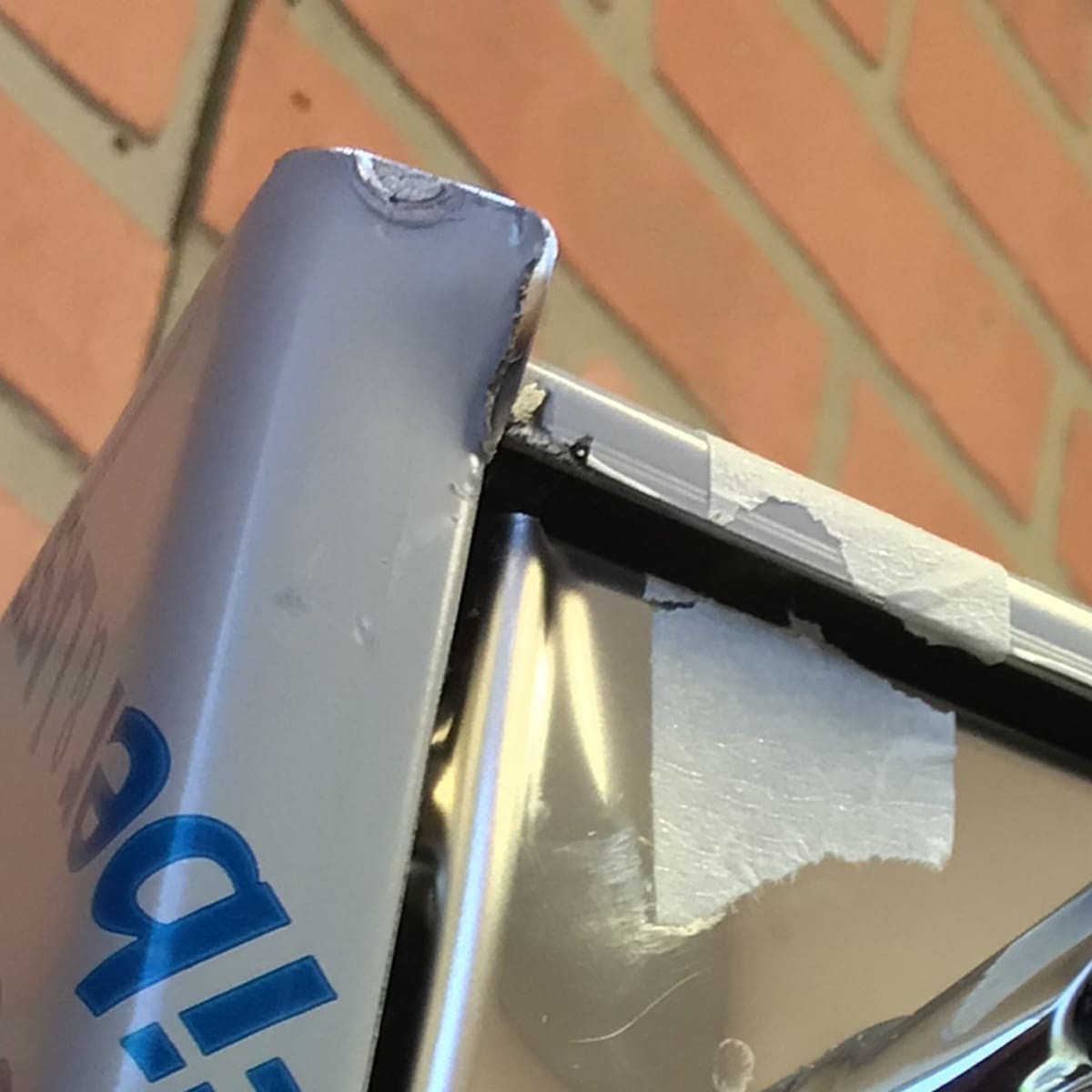 Close-up of a damaged metal sign with bent, chipped edges and peeling blue and white paint, echoing the worn finish found on a returned Bezzera Unica Espresso Machine. A blurred brick wall is visible in the background. © 2025 1st-line.com