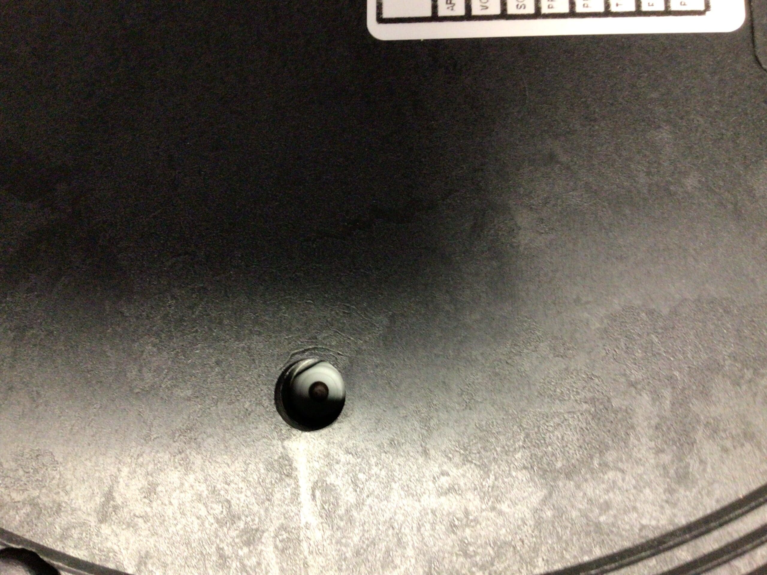 Close-up of a black surface with a small round hole near the bottom center, revealing a white and black object&mdash;possibly part of an Elektra Microcasa a Leva Safety Thermostat Reset. A white sticker with a grid and text is visible at the images top edge. &copy; 2025 1st-line.com