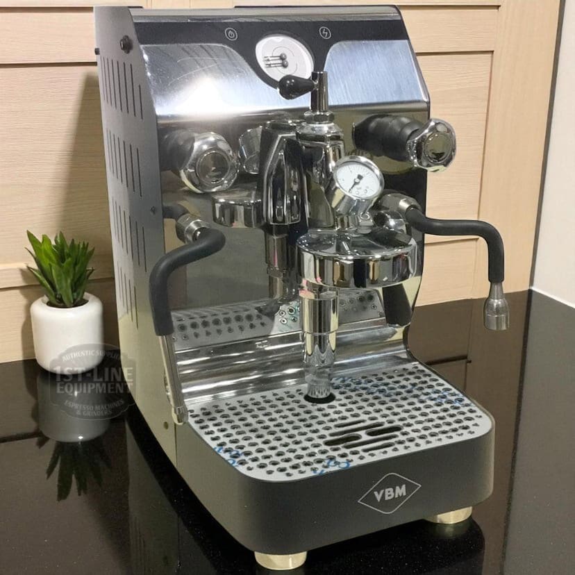 A SIDEWALK SALE VBM Domobar Super Analogic HX Espresso Machine (SHOWROOM DEMO) E011 sits on a black countertop near a small potted plant, with gauges, levers, and steam wands; a wooden cabinet is in the background. © 2025 1st-line.com