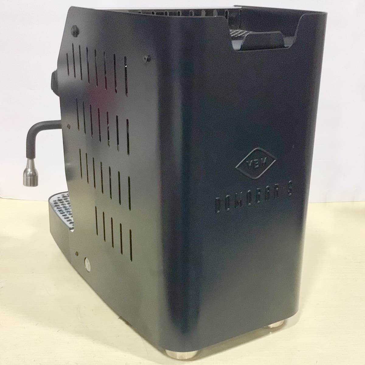 A black espresso machine with a steam wand on the side, ventilation slots, and the Dominator S logo embossed on its side panel, placed on a light-colored surface. © 2025 1st-line.com