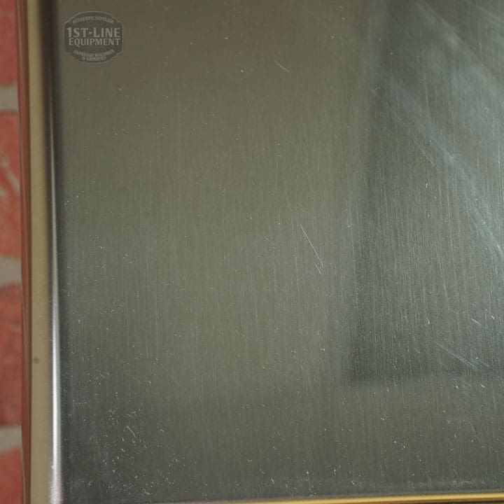 A close-up of a brushed stainless steel surface with faint scratches, a small dent & scratch, and the 1st-Line Equipment logo. The reflection of the Lelit 1000156 External Water Tank Assembly (E112) is visible on the right side. © 2025 1st-line.com