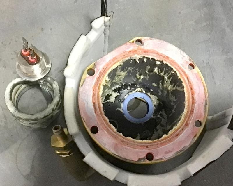 A close-up of a disassembled Rancilio Silvia Pro X coffee boiler reveals a circular component with a blue central hole, worn seals, wires, a metal fitting, and a coiled cable on a gray surface—showcasing possible limescale buildup. © 2025 1st-line.com