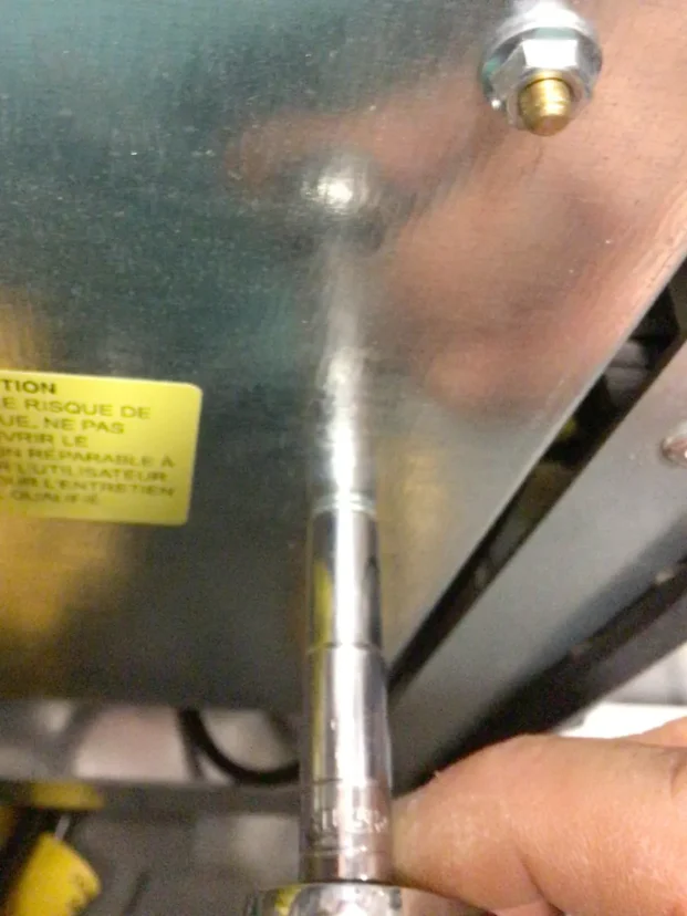 A close-up of a hand using a socket wrench to unscrew a bolt from a Carimali Bubble metal panel, with a yellow warning label visible on the left side—part of the process to swap triac components. © 2025 1st-line.com