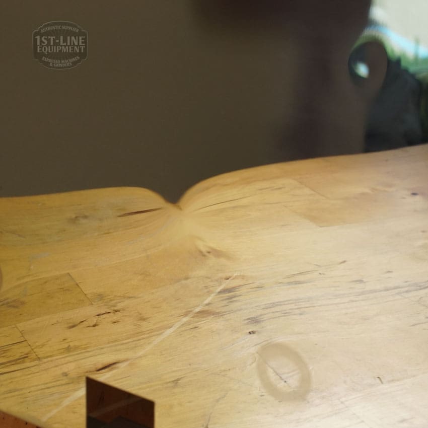 A close-up of a Lelit 1400160 Back Body Panel with noticeable dents and scratches. The faint 1st-Line Equipment logo appears in the top left corner, while a blurred green object can be seen in the background. © 2025 1st-line.com