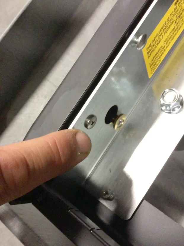 A close-up of a persons finger pointing at a metal panel with screws and holes, possibly on an electronic or mechanical device like the Carimali Bubble. A yellow warning label is partially visible at the top right. © 2025 1st-line.com