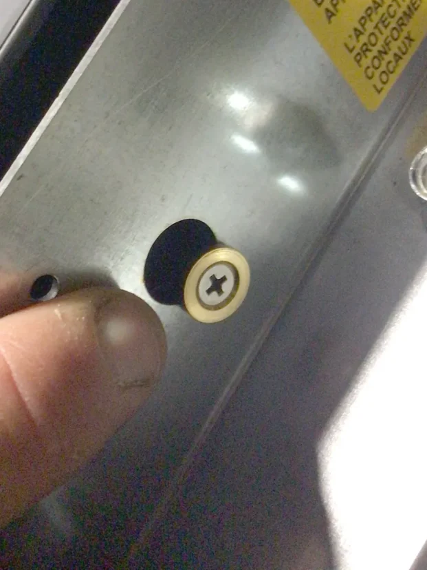 A close-up of a person’s fingers holding a metal surface near a Phillips-head screw partially sunk into the metal, possibly working on a Carimali Bubble. A yellow warning label is visible in the top right corner. © 2025 1st-line.com