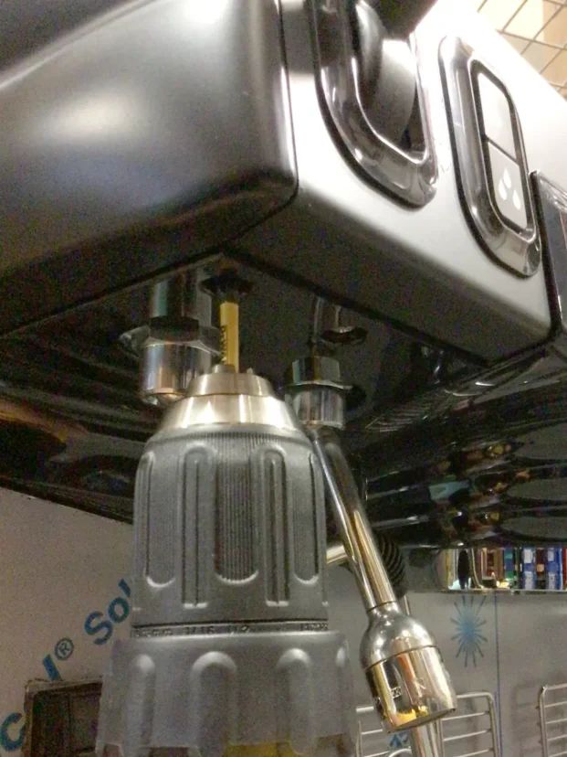 A close-up of a power drill being used to unscrew a bolt from the bottom of a black Carimali Bubble appliance, possibly a coffee machine, with reflections visible on its surface during a Swap Triac repair. © 2025 1st-line.com