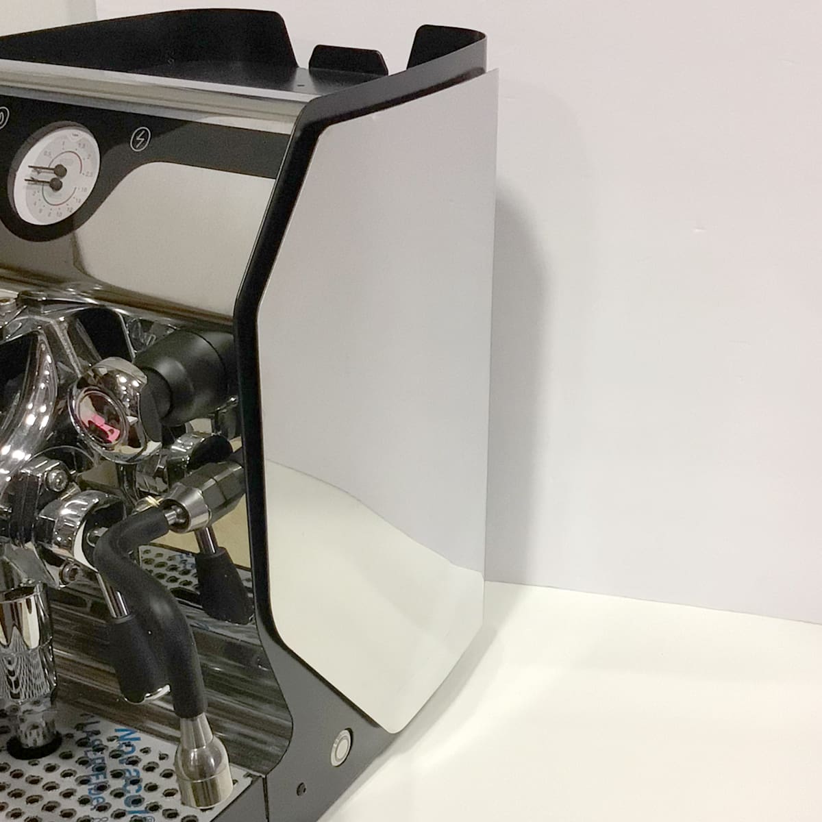 A close-up of a shiny, stainless steel espresso machine with a pressure gauge, steam wand, and drip tray, positioned on a white countertop against a plain white wall. © 2025 1st-line.com