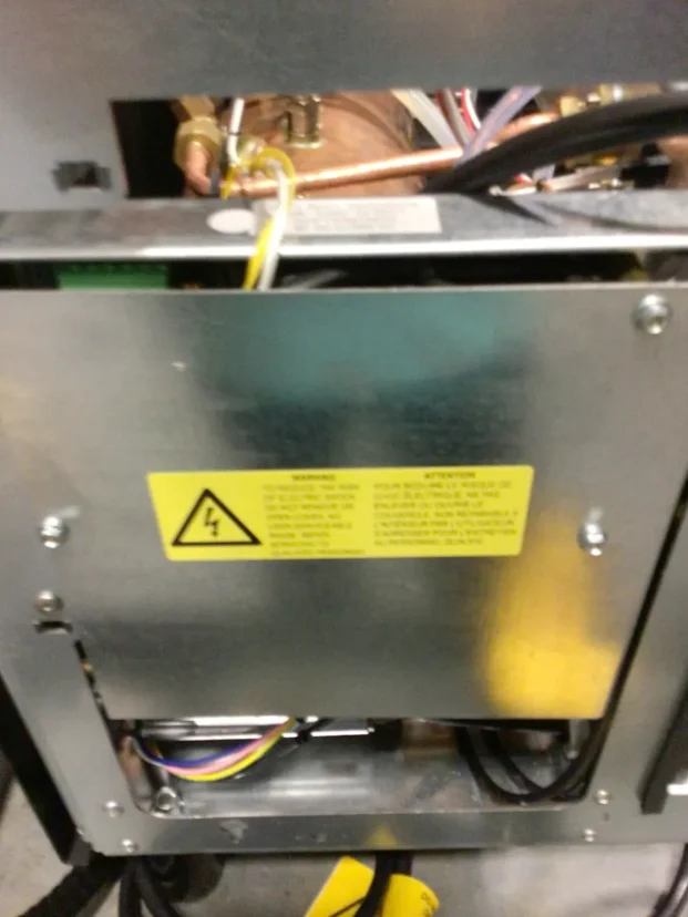 A close-up of an open metal electrical panel with wires inside, a yellow caution label warning of electrical hazards, and various colored cables visible near the bottom. © 2025 1st-line.com