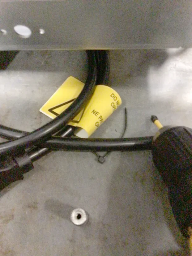 A close-up of black power cables, a yellow warning label, and a black-handled screwdriver lying on a metal surface. A silver screw is also visible nearby. © 2025 1st-line.com