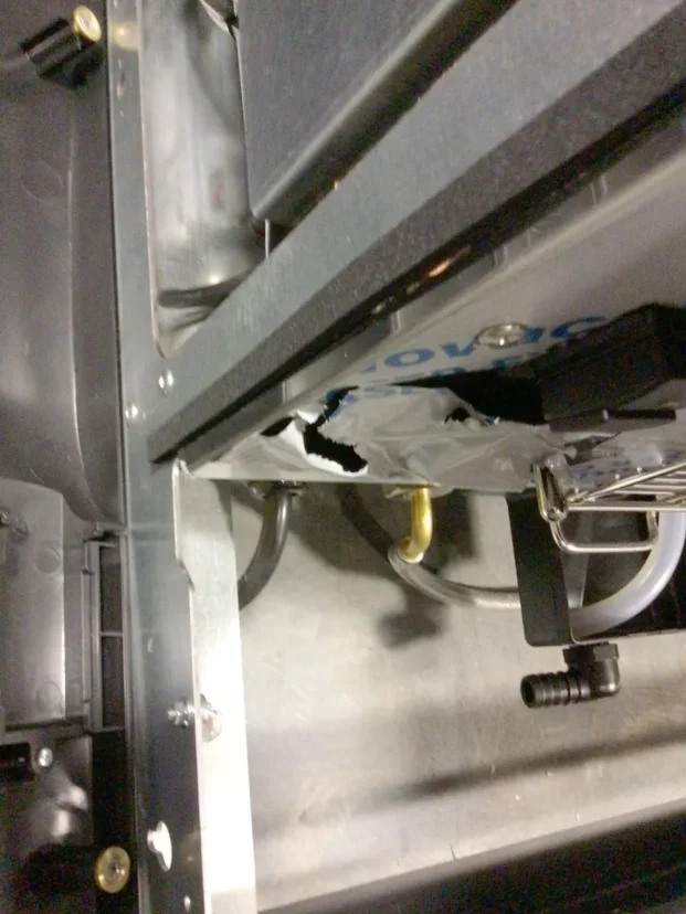 A close-up view of a damaged plastic bag inside a metal-framed compartment of a Carimali Bubble, showing a large tear and tubing nearby. Various mechanical parts, connectors, and the Swap Triac are visible around the area. © 2025 1st-line.com