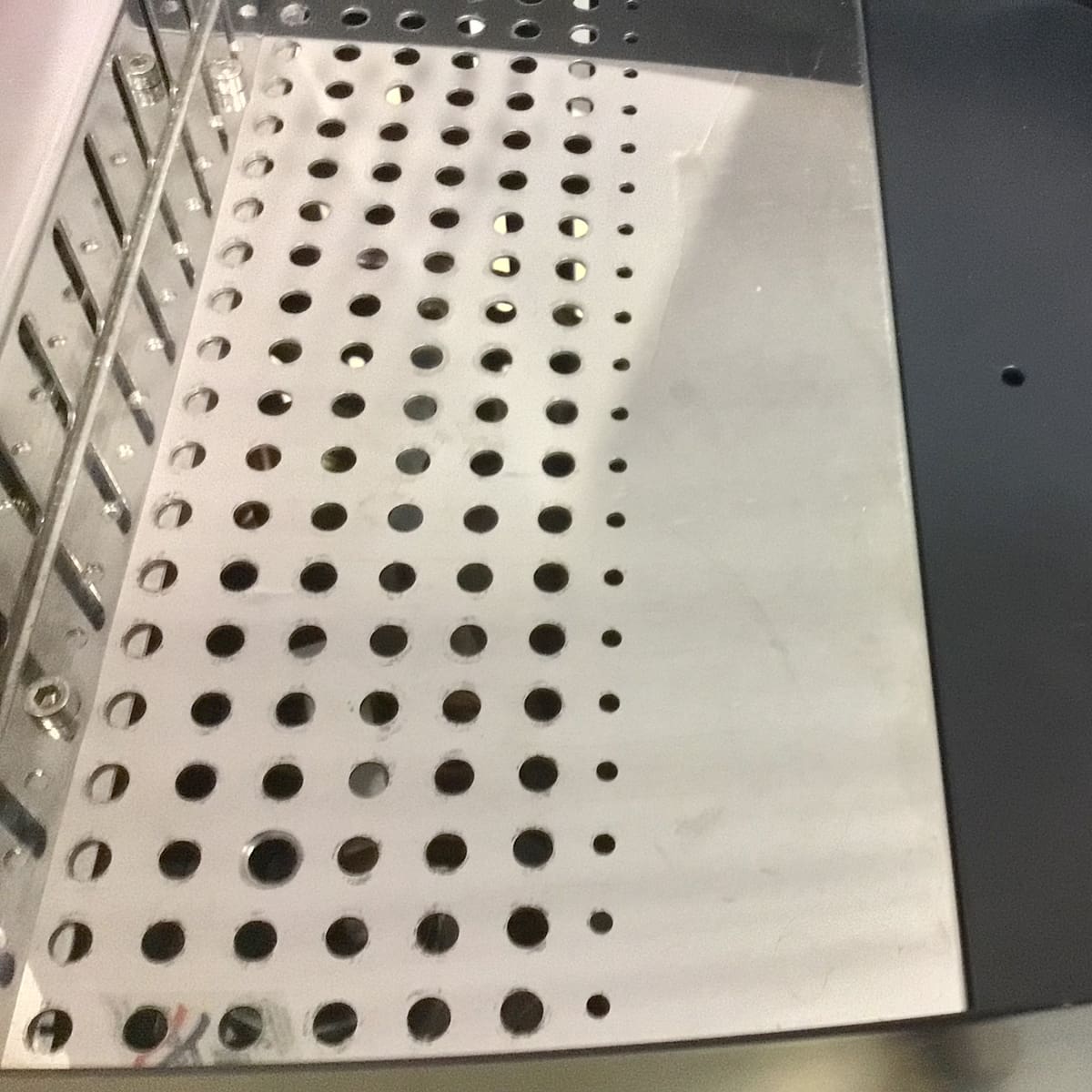 A close-up view of a metallic surface with a grid of circular holes, reflecting light. The holes are arranged in neat rows, and part of a dark edge is visible on the right side. © 2025 1st-line.com