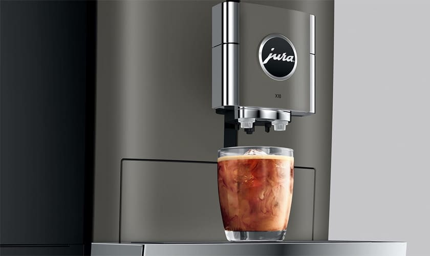 A glass of iced coffee sits under the sleek, metallic Jura X10 Super-Automatic Espresso Machine as fresh coffee pours over ice cubes. © 2025 1st-line.com