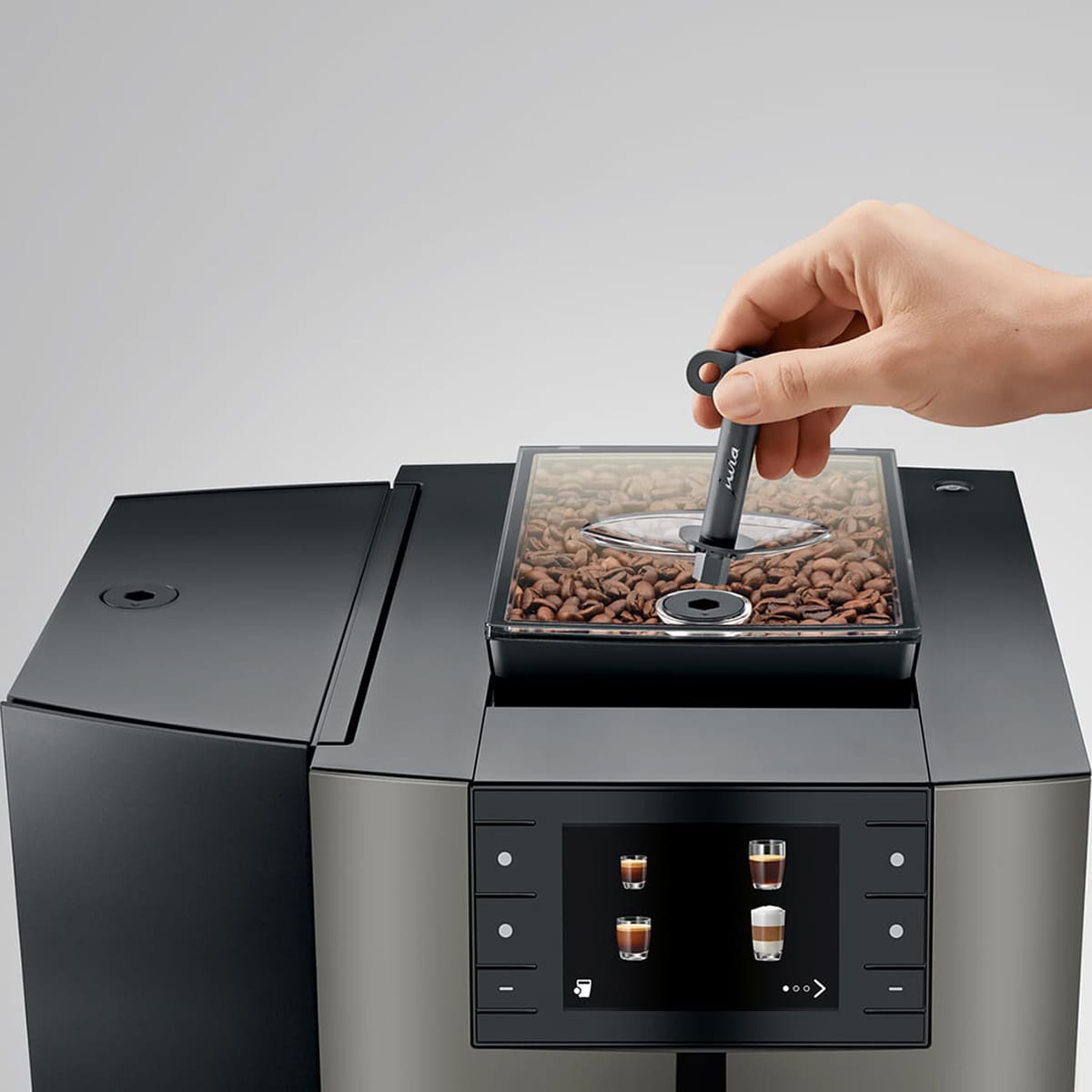 A hand uses a measuring spoon to add coffee beans into the top compartment of the Jura X10 Super-Automatic Espresso Machine, which features a digital display showing images of various coffee drinks. © 2025 1st-line.com