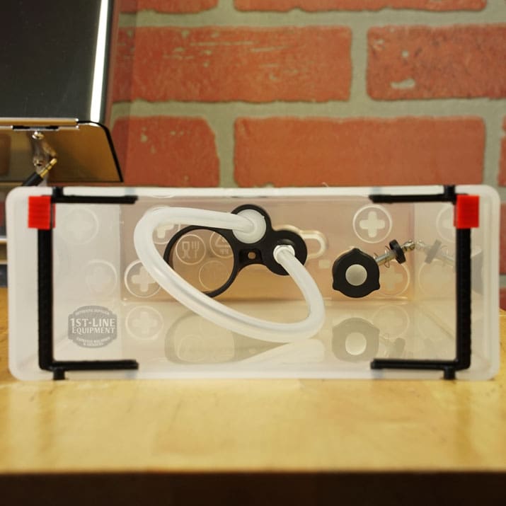 A Lelit 1000156 External Water Tank Assembly - Small Dent & Scratch (E112), featuring white tubing, black fittings, and black/red clips in a clear rectangular box, rests on wood with a brick wall backdrop—perfect for water tank setups. © 2025 1st-line.com