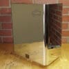 A Lelit 1400160 Back Body Panel with dents, made of shiny stainless steel, is positioned on a wooden surface before a red brick wall, reflecting the bricks on its polished finish. © 2025 1st-line.com