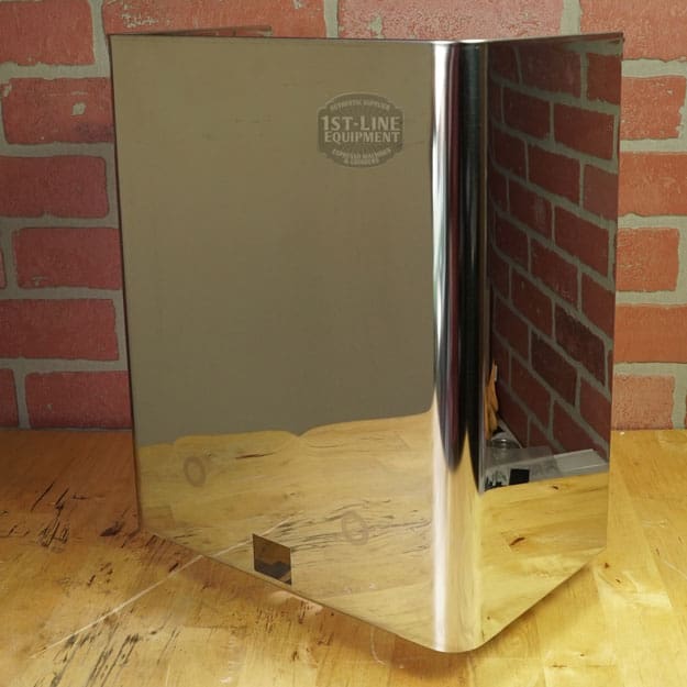 A Lelit 1400160 Back Body Panel with dents, made of shiny stainless steel, is positioned on a wooden surface before a red brick wall, reflecting the bricks on its polished finish. © 2025 1st-line.com