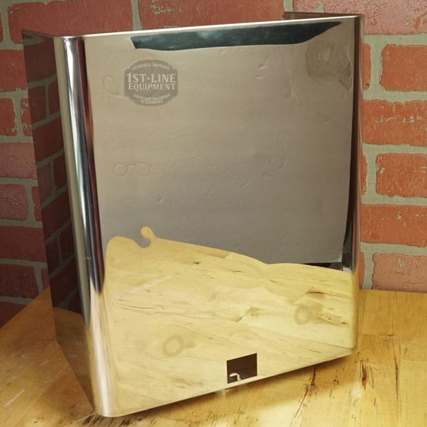 A Lelit 1400160 Back Body Panel with dents sits on a wooden surface against a red brick wall, its shiny, reflective metal finish capturing the tabletop and surroundings. © 2025 1st-line.com