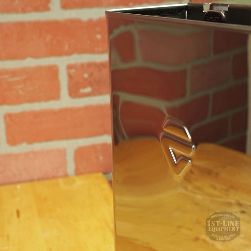 A shiny, dark Lelit 1000156 External Water Tank Assembly - Small Dent & Scratch (E112) sits on a wooden surface before a red brick wall, displaying a geometric logo and reflections on its surface. © 2025 1st-line.com