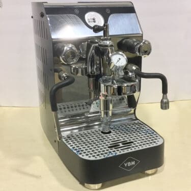 A shiny VBM espresso machine with two black handles, a pressure gauge, and a metal drip tray, set on a light-colored surface. The machine has a polished metal and black finish. © 2025 1st-line.com