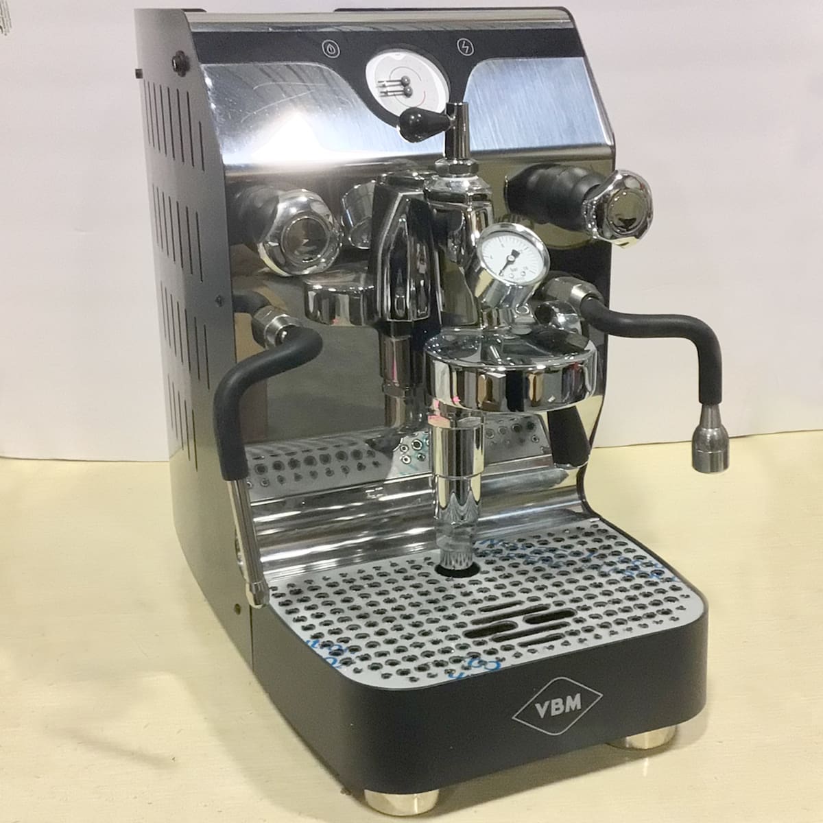 A shiny VBM espresso machine with two black handles, a pressure gauge, and a metal drip tray, set on a light-colored surface. The machine has a polished metal and black finish. © 2025 1st-line.com