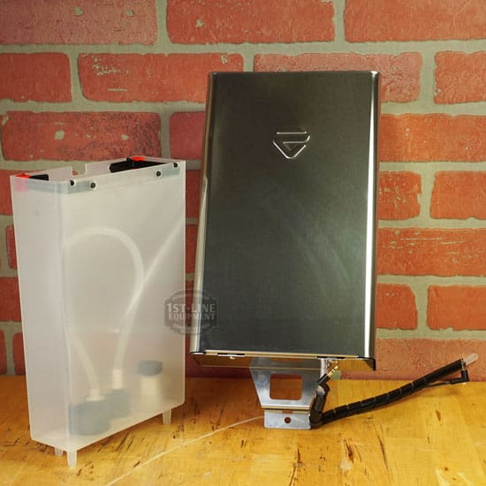 A sleek metal battery pack and a translucent plastic battery case with visible wires sit beside the Lelit 1000156 External Water Tank Assembly - Small Dent & Scratch (E112) on a wooden surface against a red brick wall background. © 2025 1st-line.com