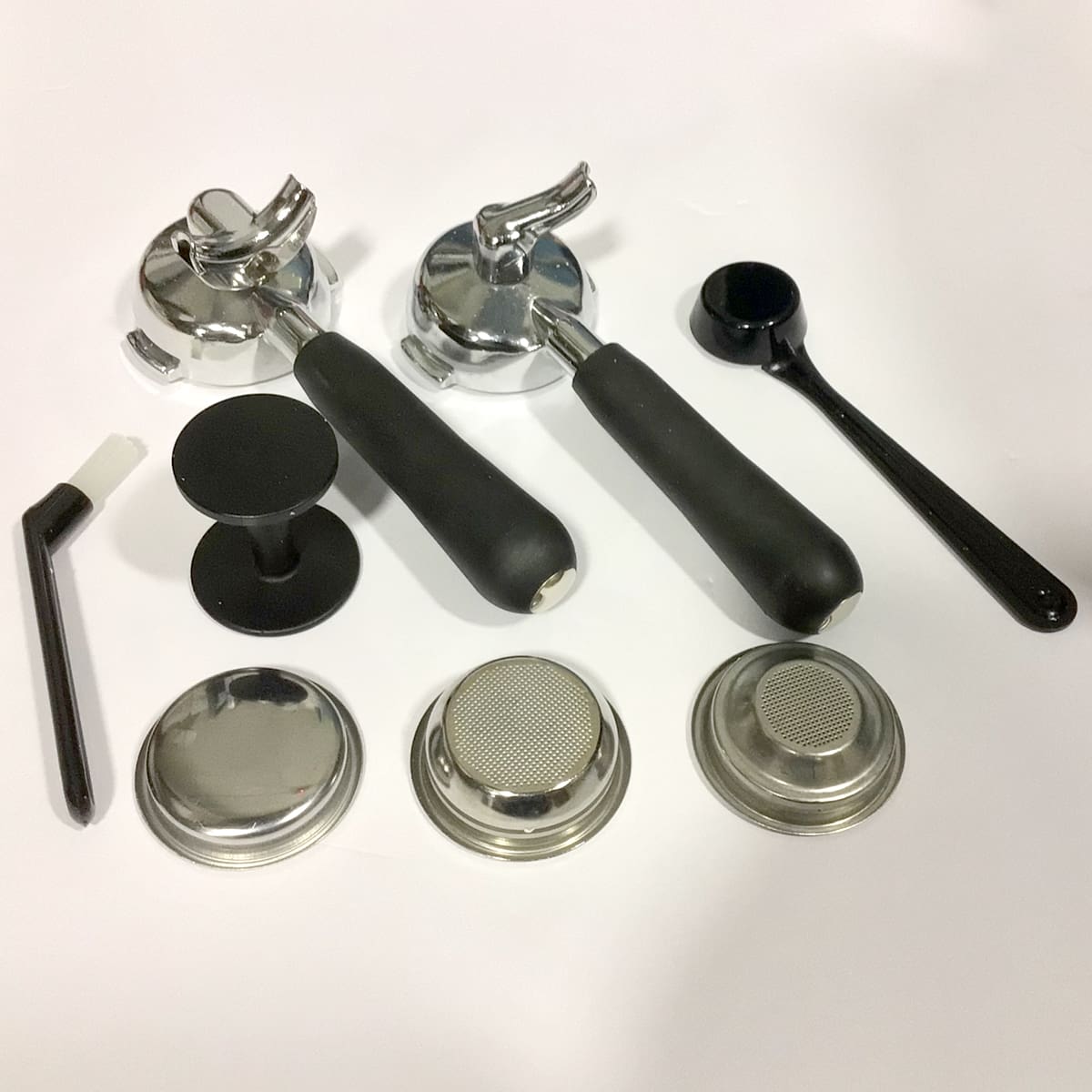 Assorted coffee machine accessories, including two portafilters, a black tamper, filter baskets, a dosing spoon, a small cleaning brush, and a metal mesh filter, all arranged on a white surface. © 2025 1st-line.com