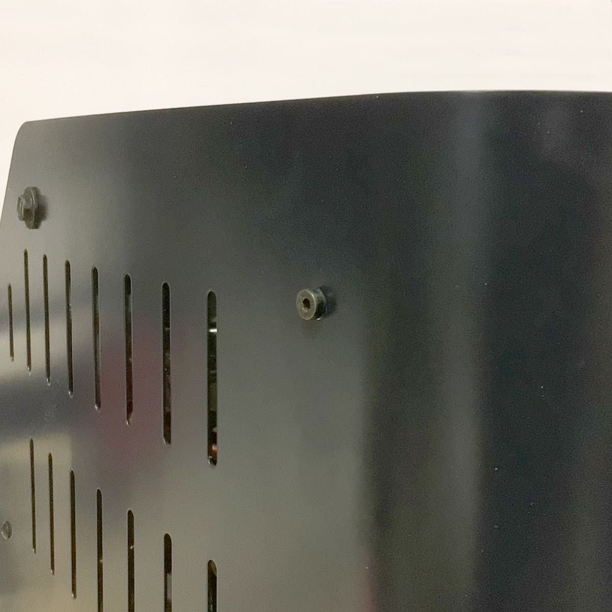 Close-up of a black metal panel with vertical ventilation slots and screws. The background is out of focus and appears to be a light-colored wall. © 2025 1st-line.com