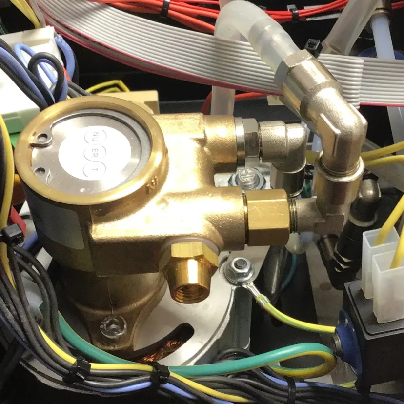 Close-up of a brass water pump with pipes, hoses, and electrical wires inside the Nurri R-Type DB Espresso Machine, revealing part of its intricate electronic internal components. © 2025 1st-line.com